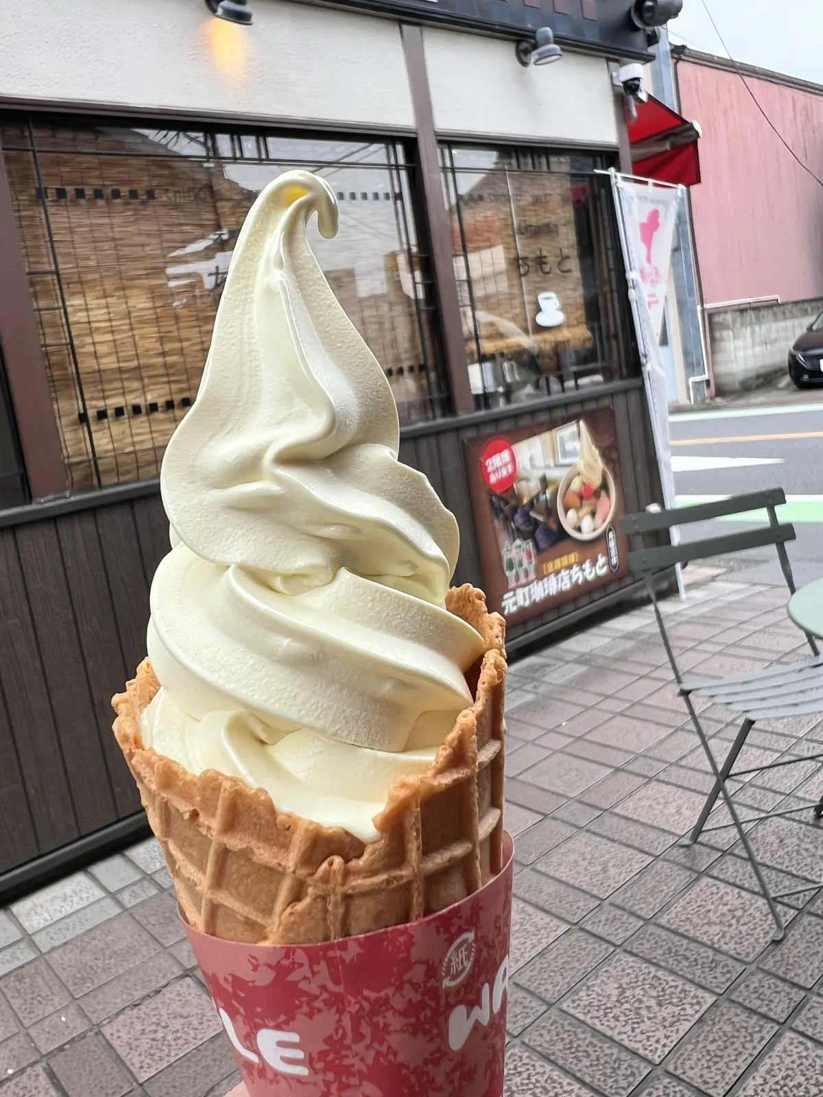 コーンに巻かれた淡い黄色の芋ソフトクリーム。背景は店の外観。