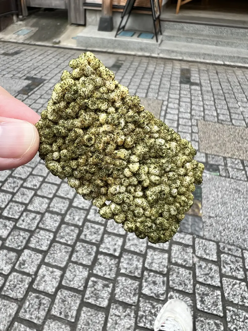 緑の煎茶塩がまぶされたおこげ揚餅を手に持っているところ。足元は石畳。